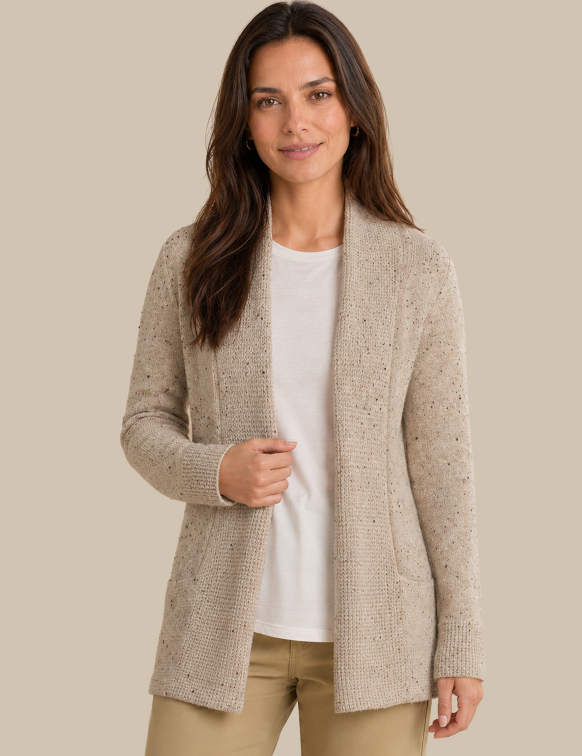 A woman modeling a light beige open-front knit cardigan over a white top and tan pants, photographed against a plain warm beige studio background.