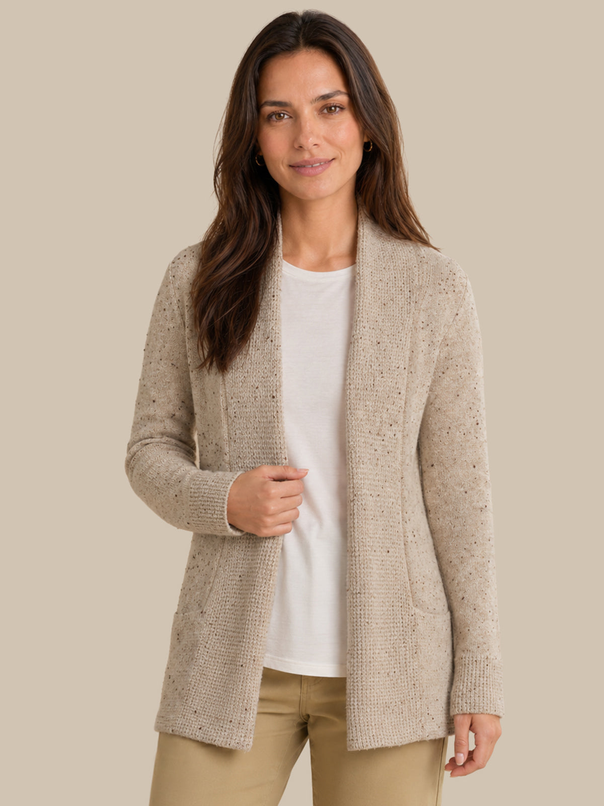 A woman modeling a light beige open-front knit cardigan over a white top and tan pants, photographed against a plain warm beige studio background.