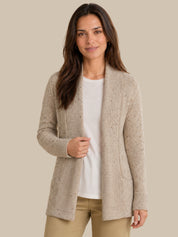 A woman modeling a light beige open-front knit cardigan over a white top and tan pants, photographed against a plain warm beige studio background.
