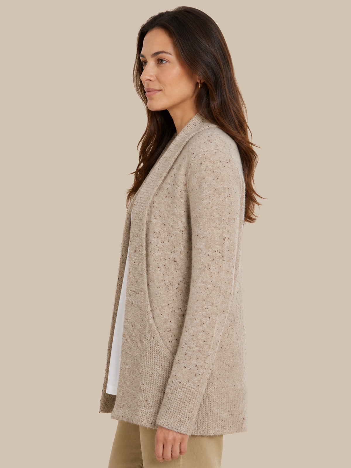 A woman shown in side profile wearing a light beige textured knit cardigan over a white top and khaki pants, set against a solid beige studio background.