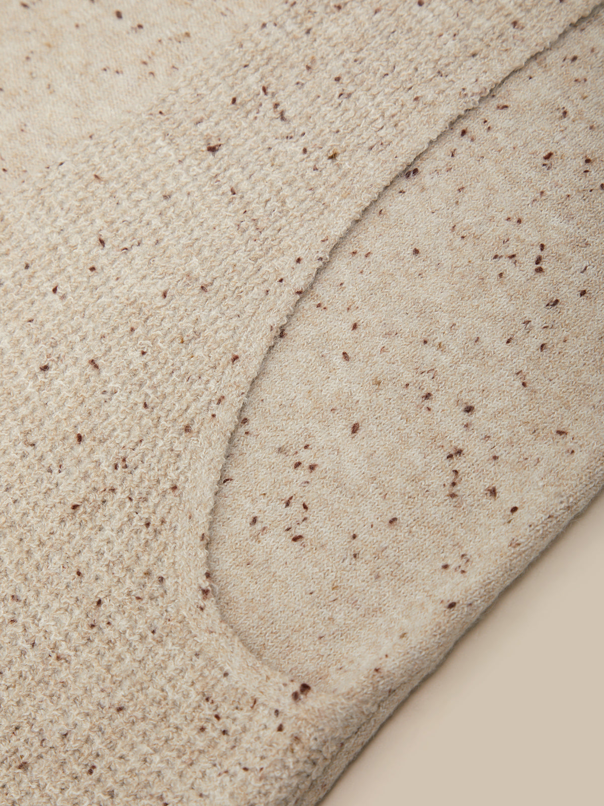 Close-up of a beige knit fabric with small brown speckles, showing a curved seam and texture detail.