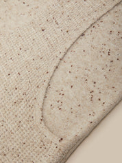 Close-up of a beige knit fabric with small brown speckles, showing a curved seam and texture detail.