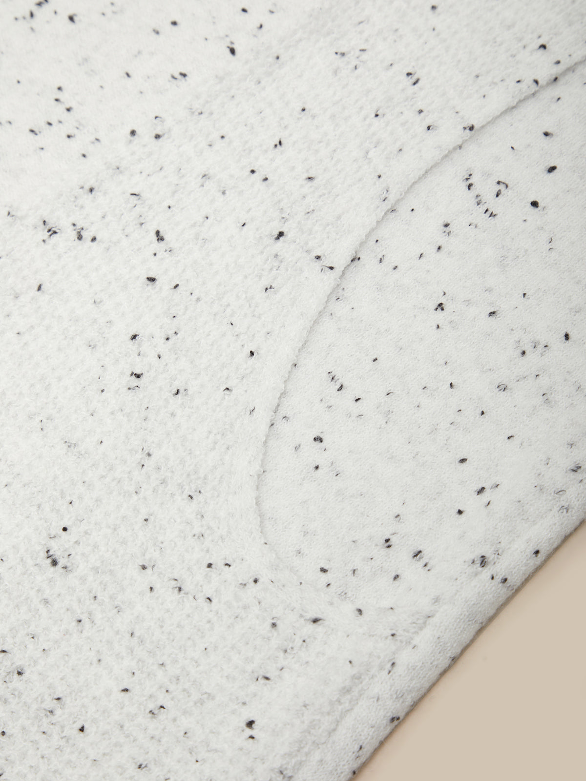 Close-up of a white textured fabric with small black speckles and a curved seam, shown against a light beige background.