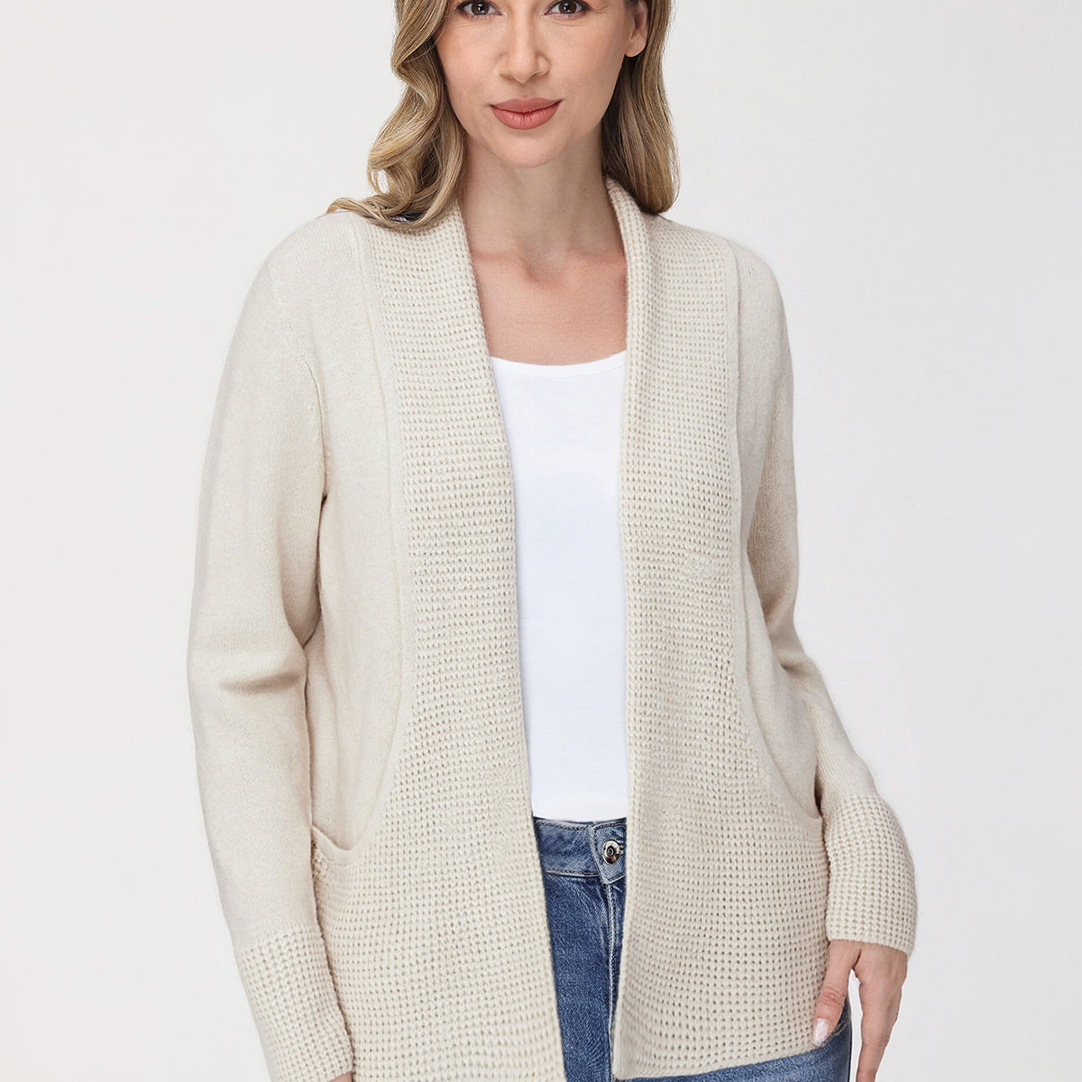 Woman wearing a beige cardigan over a white shirt and blue jeans on a white background