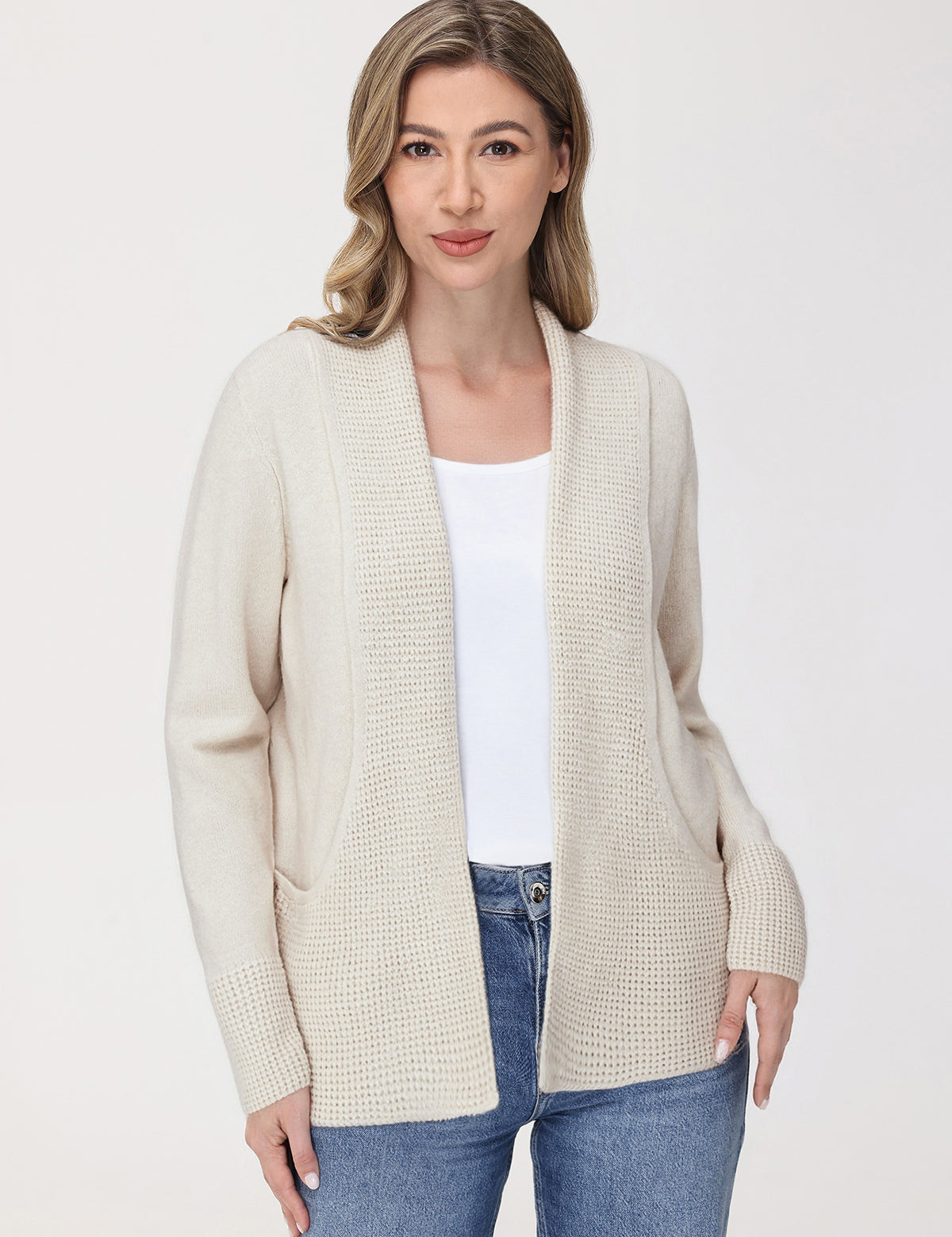 Woman wearing a beige cardigan over a white shirt and blue jeans on a white background