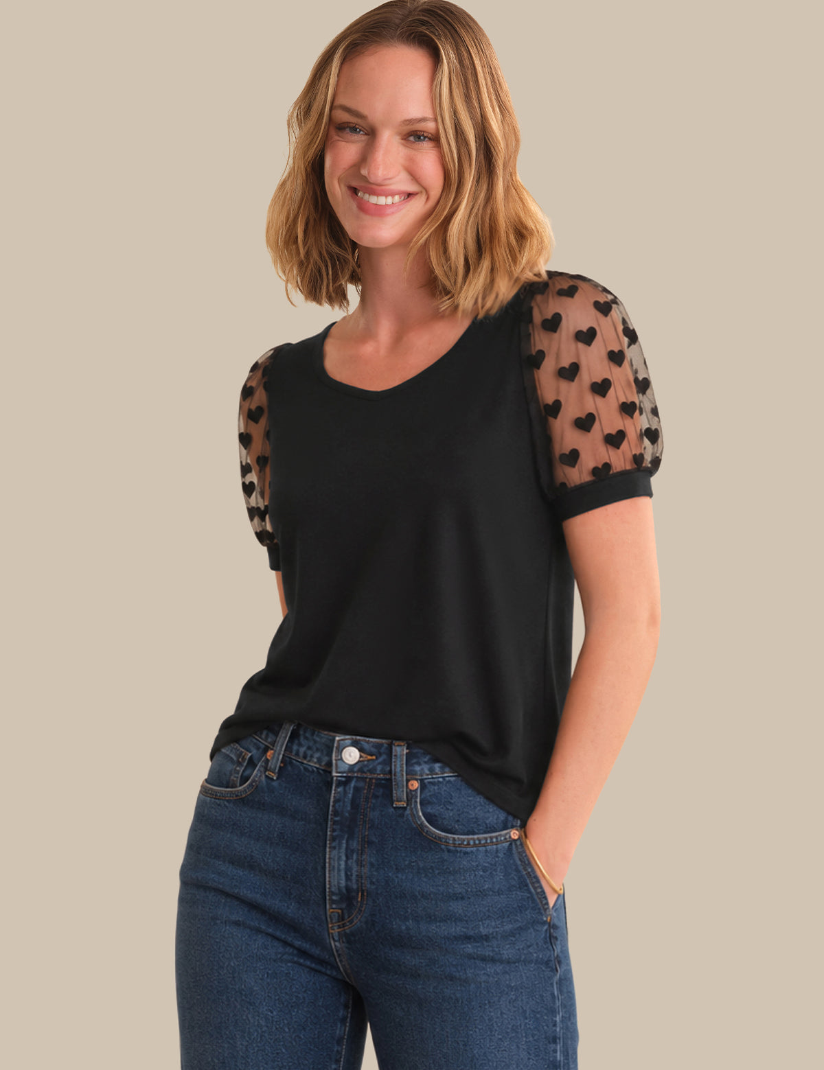 A woman wearing a black short-sleeve top with sheer patterned puff sleeves and blue jeans, standing against a solid beige background.