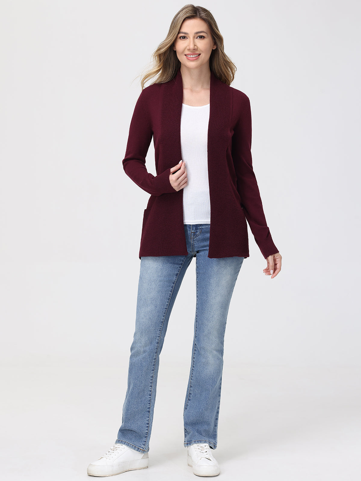 Woman wearing a burgundy cardigan, white shirt, blue jeans, and white sneakers on a white background