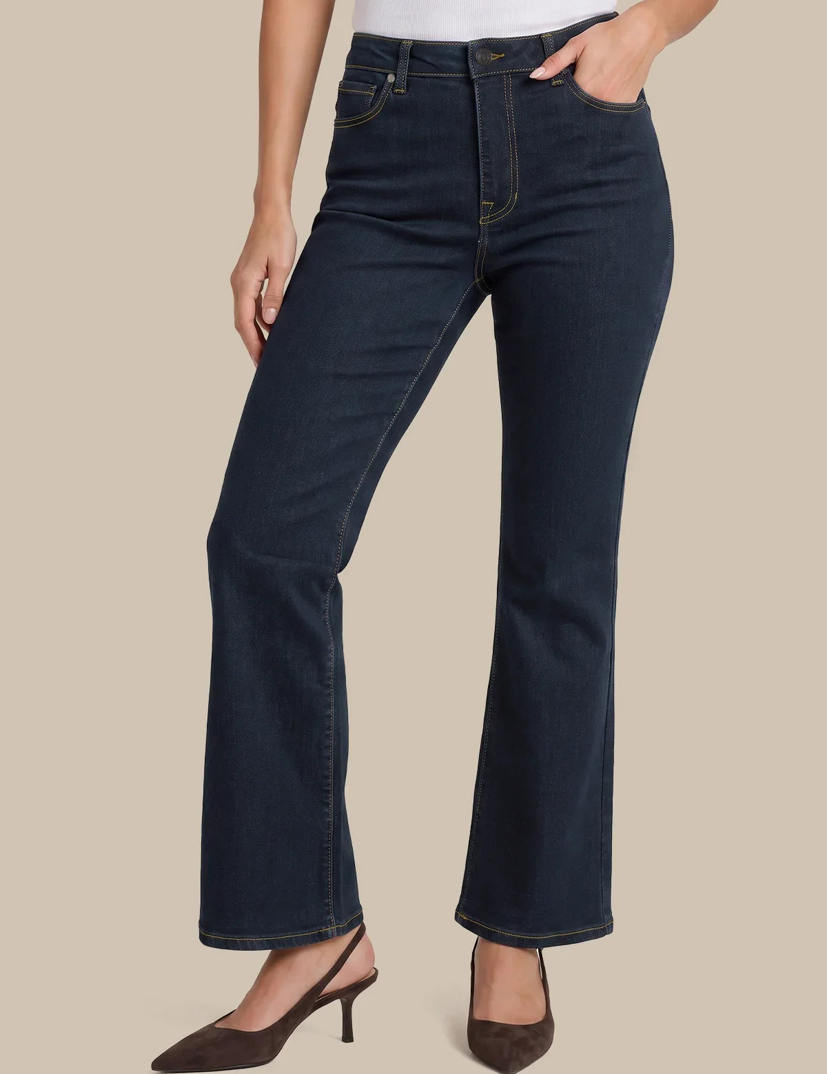 A model wearing dark blue flare jeans with a white top and brown slingback heels, shown from the waist down against a solid beige background.
