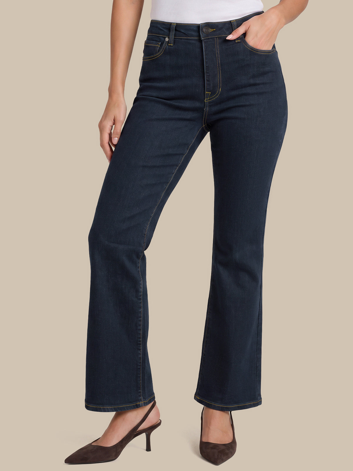 A model wearing dark blue flare jeans with a white top and brown slingback heels, shown from the waist down against a solid beige background.