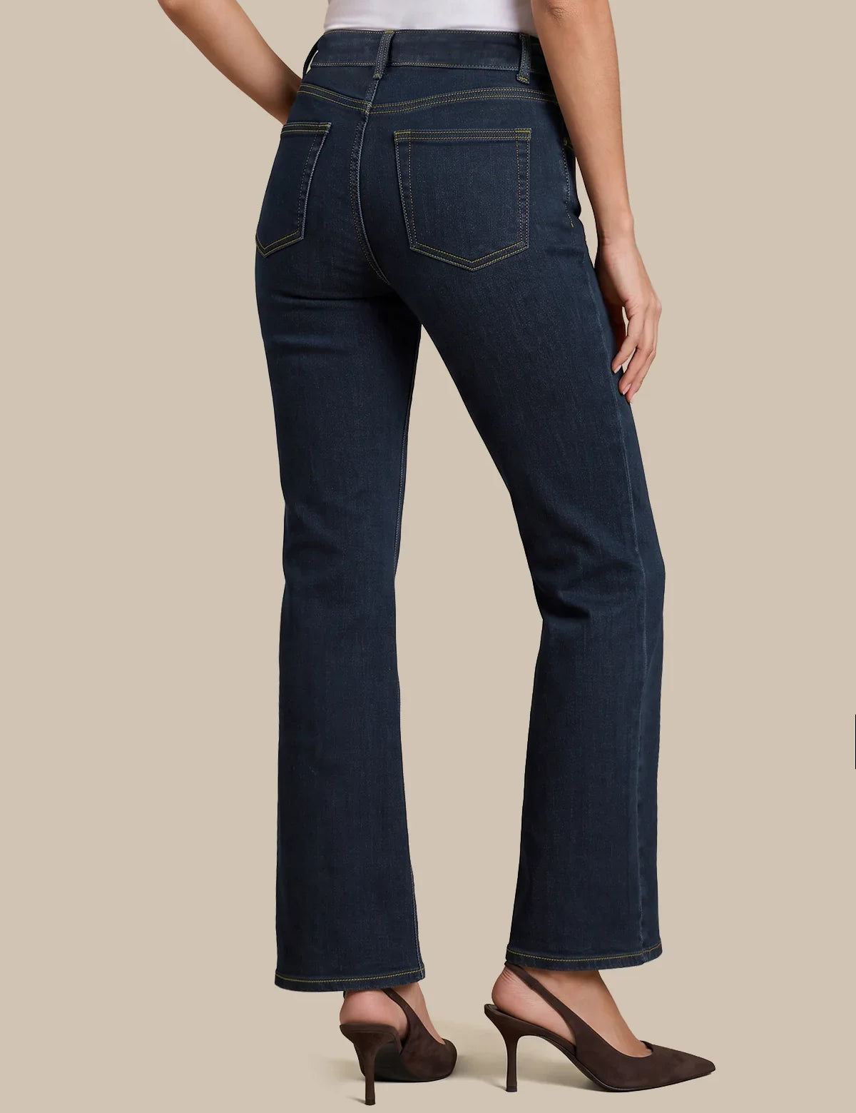 Rear view of a model wearing dark blue flare jeans with gold contrast stitching, paired with dark brown slingback heels against a solid beige background.