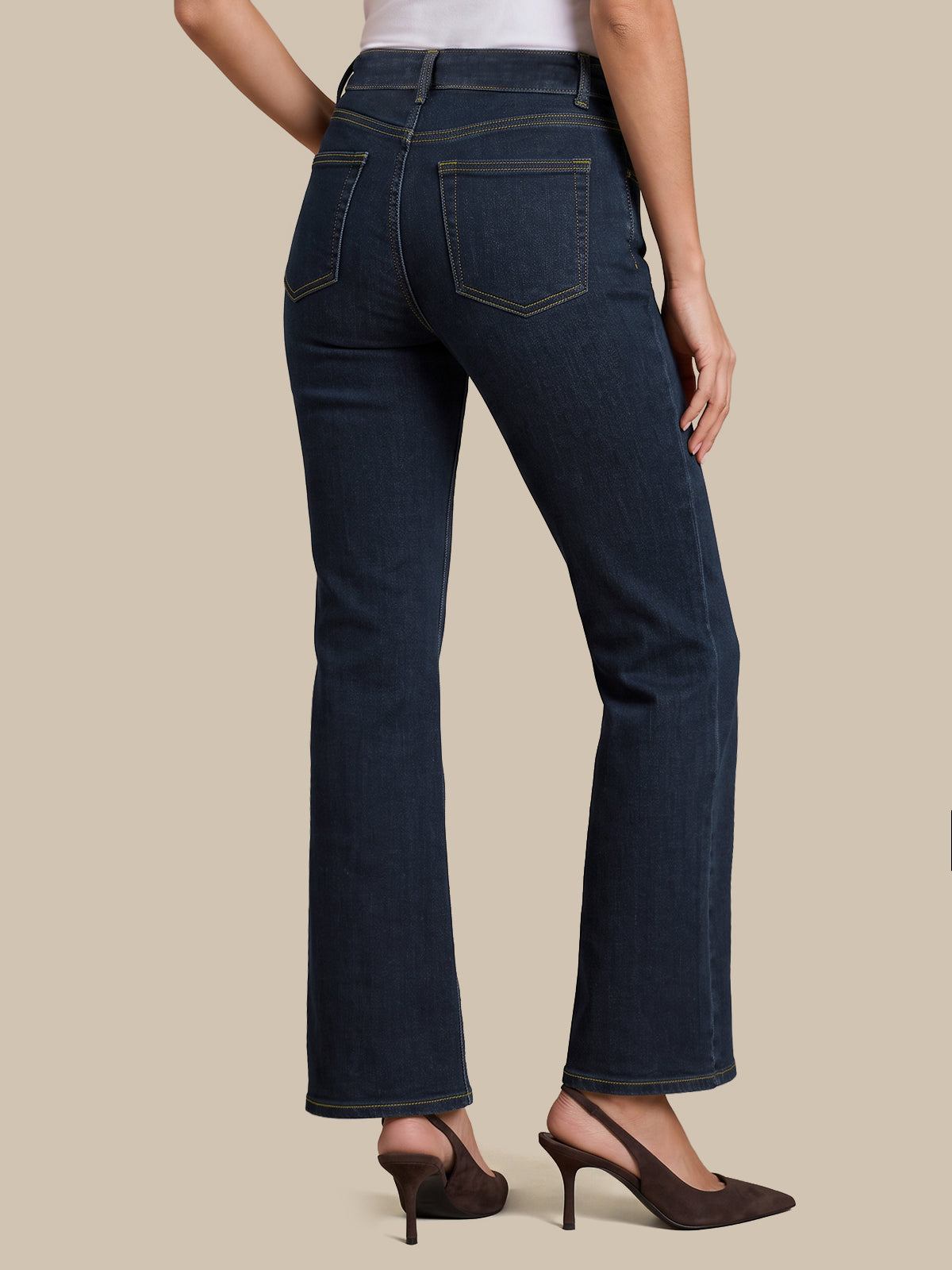 Rear view of a model wearing dark blue flare jeans with gold contrast stitching, paired with dark brown slingback heels against a solid beige background.