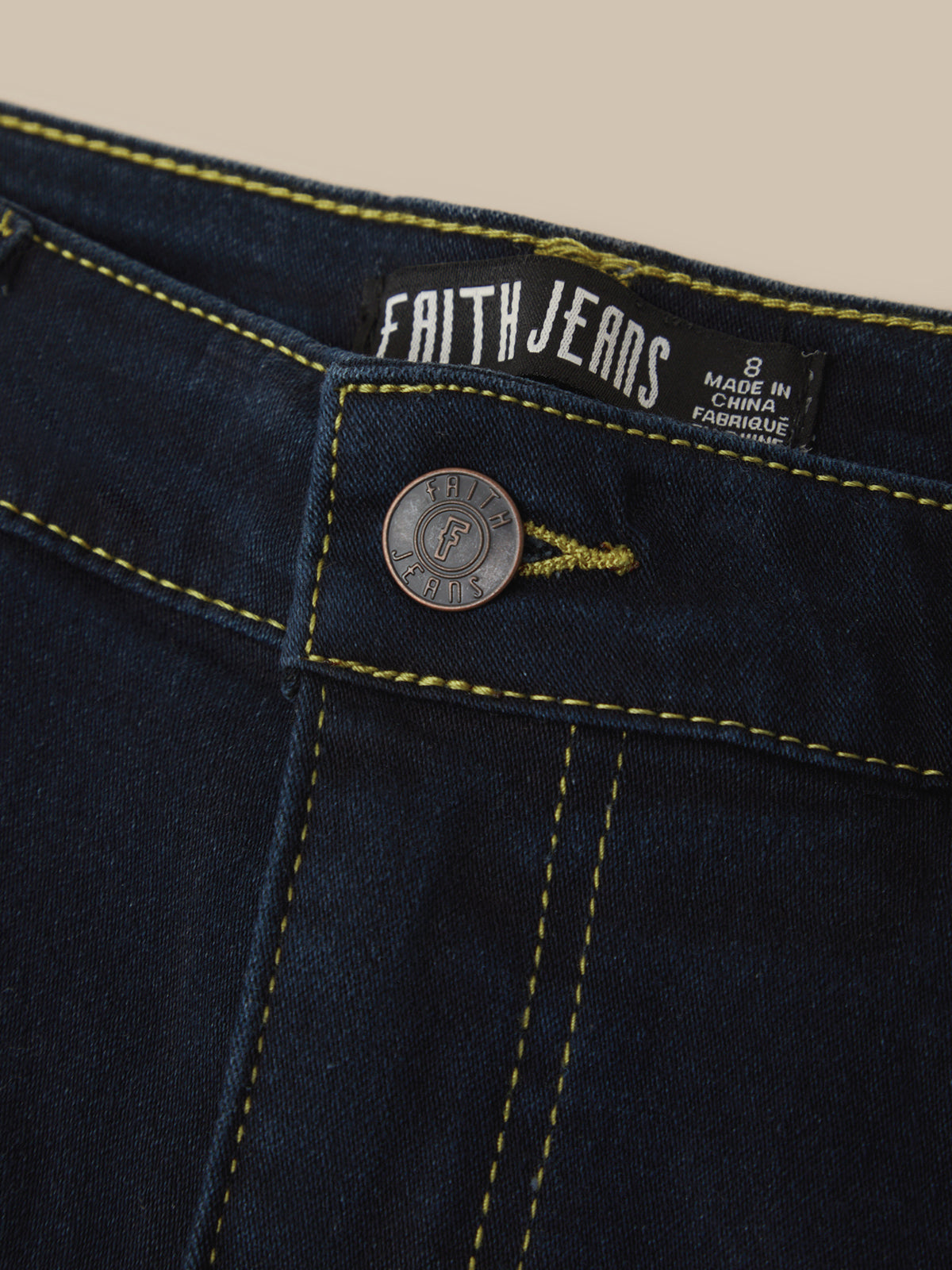 Close-up of dark indigo denim jeans showing the waistband, yellow contrast stitching, a round metal button with a Faith Jeans logo, and an interior brand label.