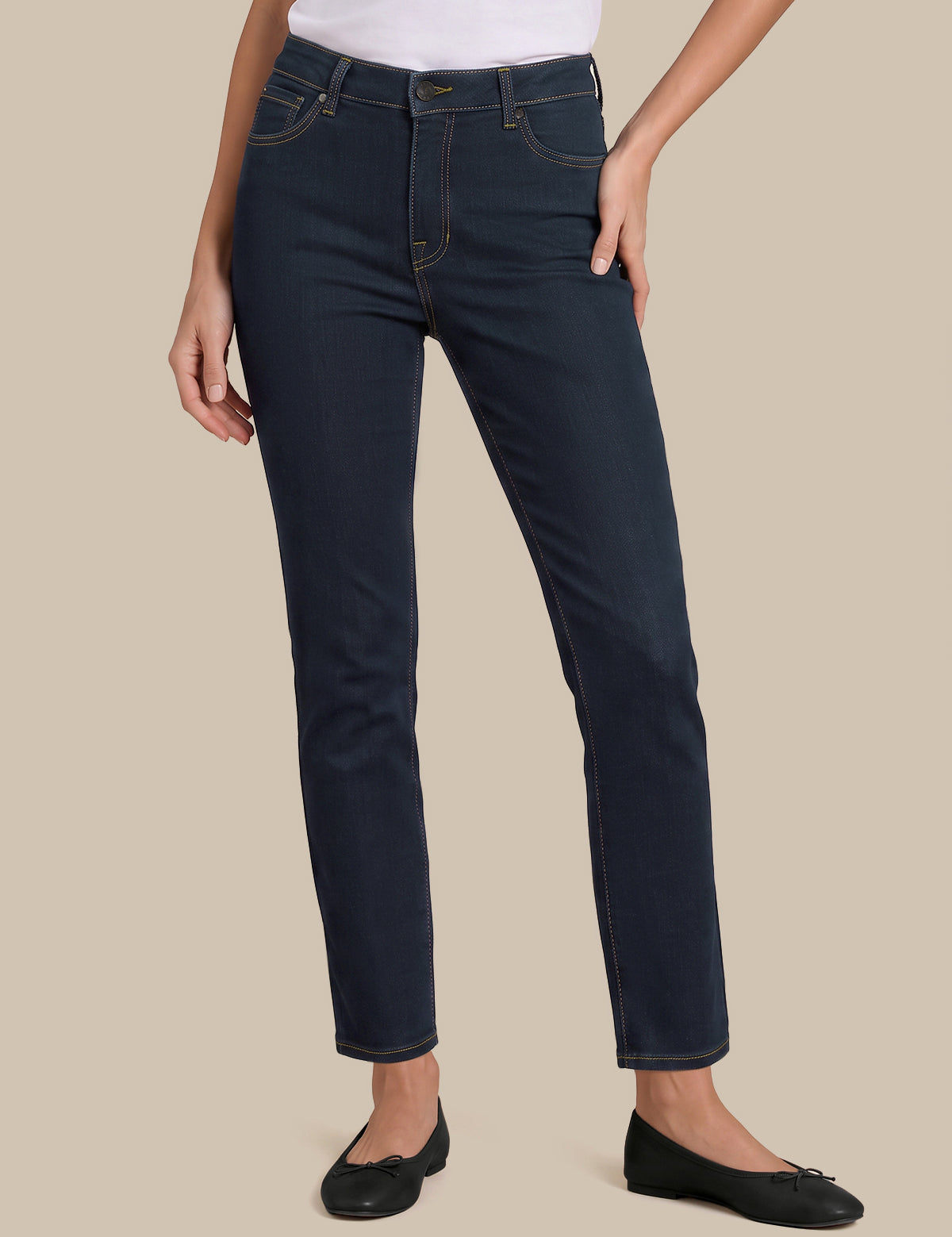 A model wearing dark navy blue slim-fit jeans, styled with a white top and black flats, standing against a solid beige background.