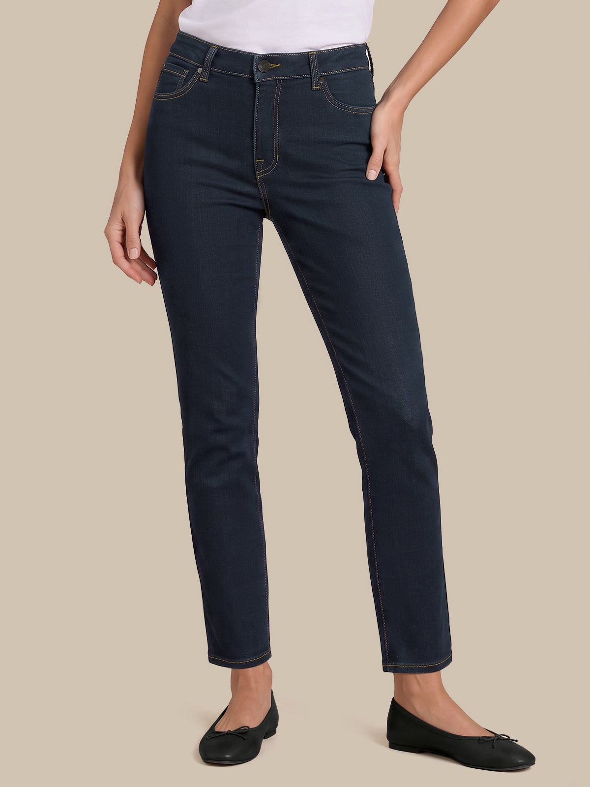 A model wearing dark navy blue slim-fit jeans, styled with a white top and black flats, standing against a solid beige background.