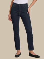A model wearing dark navy blue slim-fit jeans, styled with a white top and black flats, standing against a solid beige background.