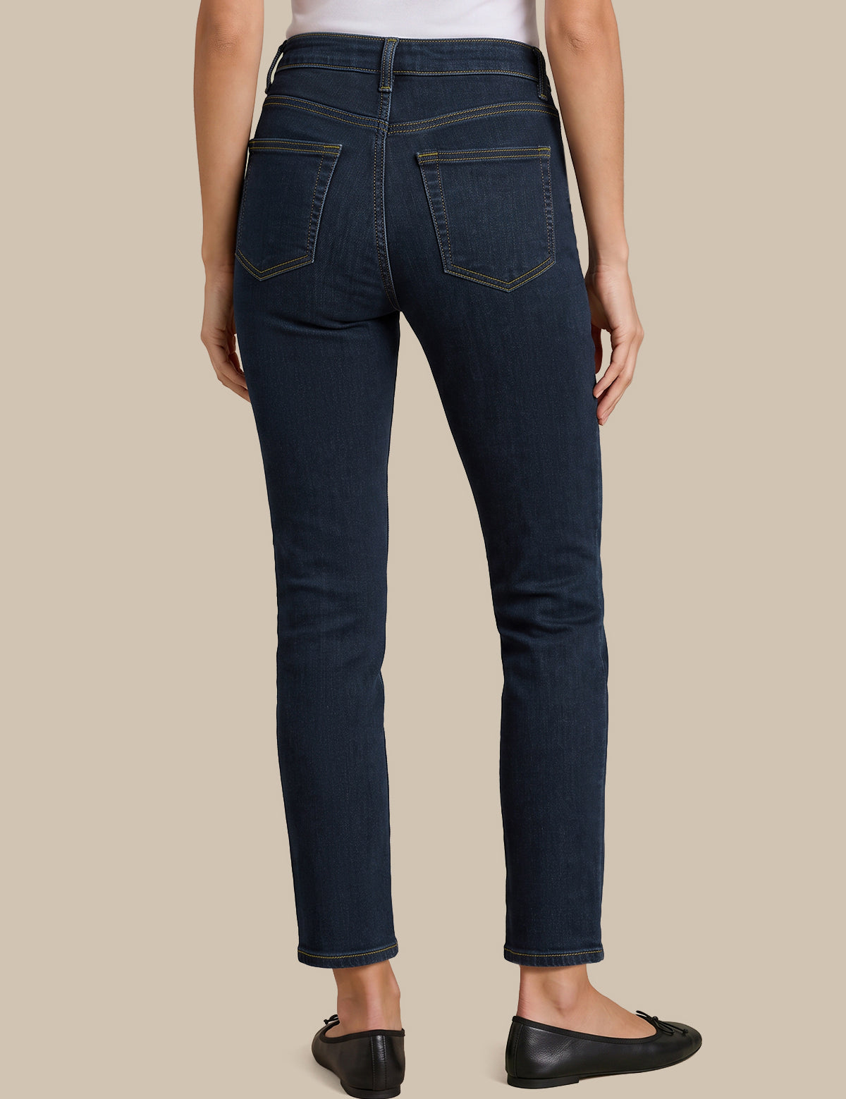 Rear view of a model wearing dark blue slim-fit jeans with gold contrast stitching, paired with a white top and black flats, against a solid beige background.