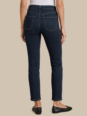 Rear view of a model wearing dark blue slim-fit jeans with gold contrast stitching, paired with a white top and black flats, against a solid beige background.