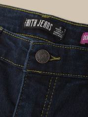 Close-up of dark blue denim jeans showing the waistband, a black Faith Jeans brand label, a round metal button, and yellow contrast stitching against a light beige background.