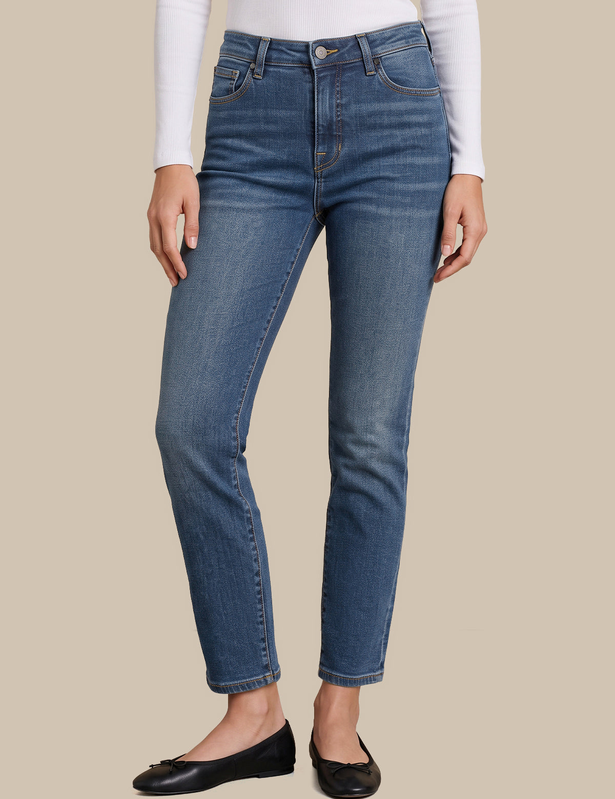 A model wearing a pair of medium-wash blue denim jeans with a straight-to-ankle leg, styled with a white long-sleeve top and black flat shoes against a solid beige background.