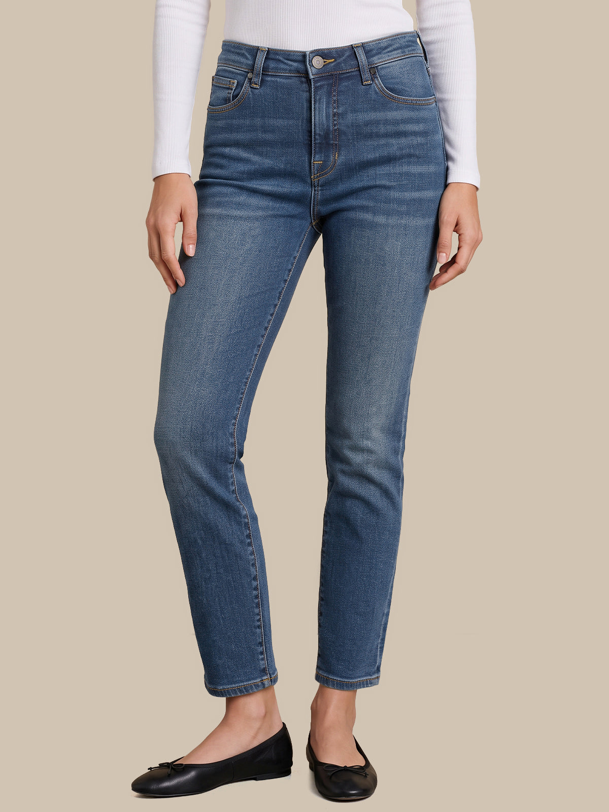 A model wearing a pair of medium-wash blue denim jeans with a straight-to-ankle leg, styled with a white long-sleeve top and black flat shoes against a solid beige background.