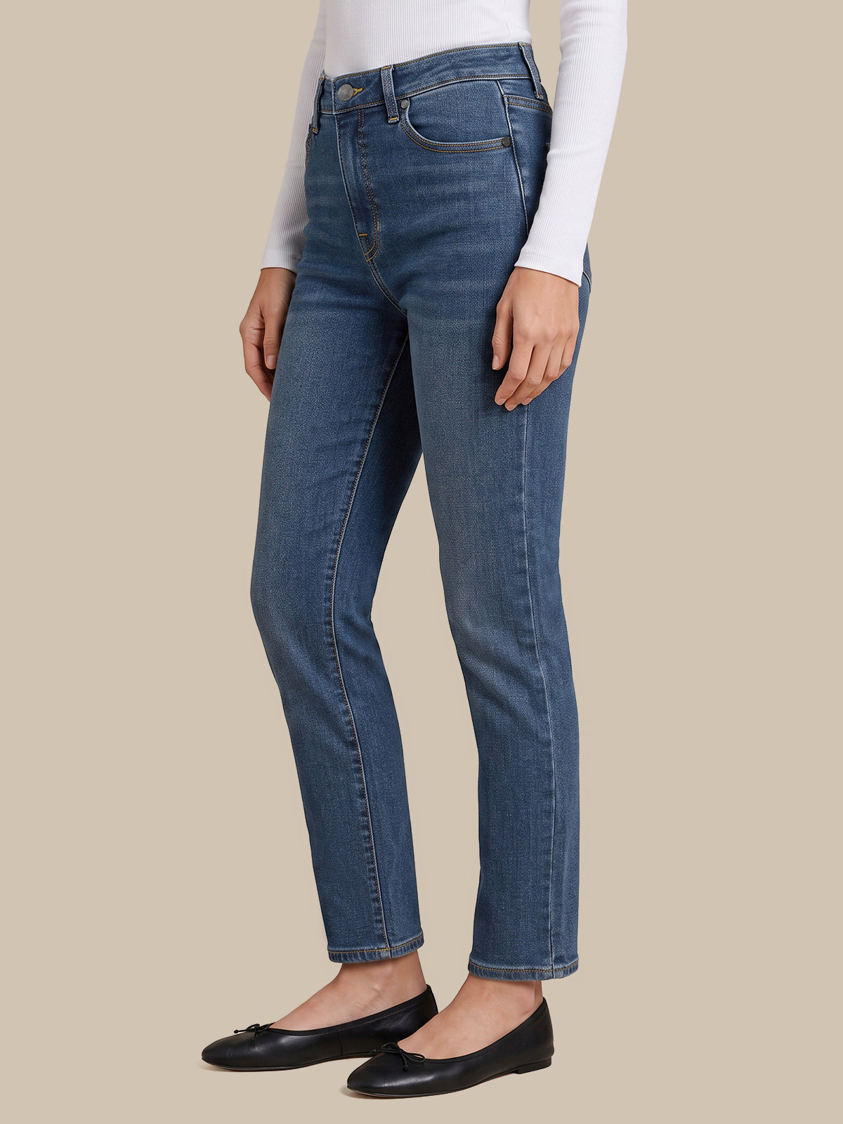 A model wearing a pair of medium-wash blue denim jeans, styled with a white long-sleeve top and black flat shoes, shown from the waist down against a solid beige background.