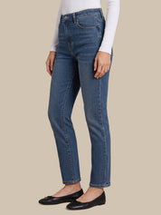 A model wearing a pair of medium-wash blue denim jeans, styled with a white long-sleeve top and black flat shoes, shown from the waist down against a solid beige background.