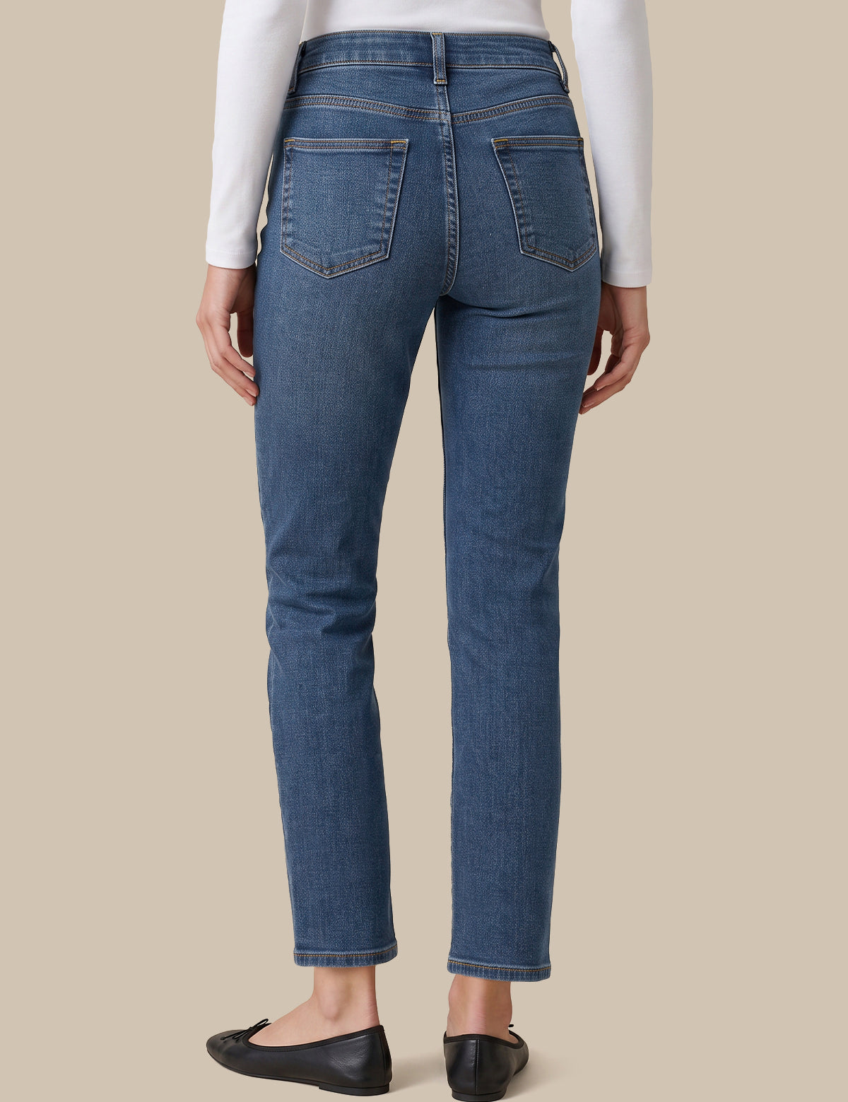 Rear view of a person wearing blue straight-leg jeans, a white long-sleeve top, and black flats against a solid beige background.