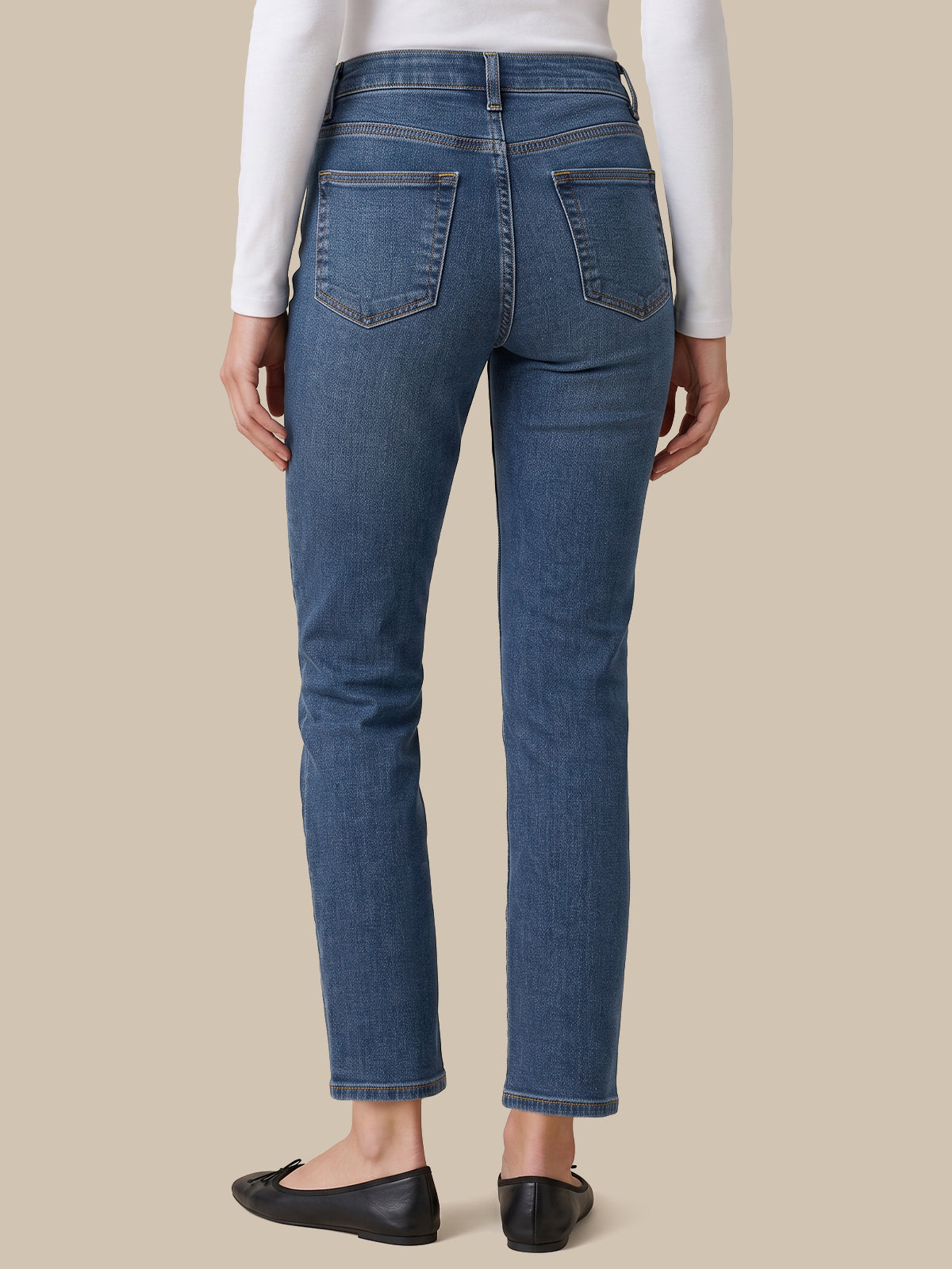 Rear view of a person wearing blue straight-leg jeans, a white long-sleeve top, and black flats against a solid beige background.