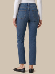 Rear view of a person wearing blue straight-leg jeans, a white long-sleeve top, and black flats against a solid beige background.