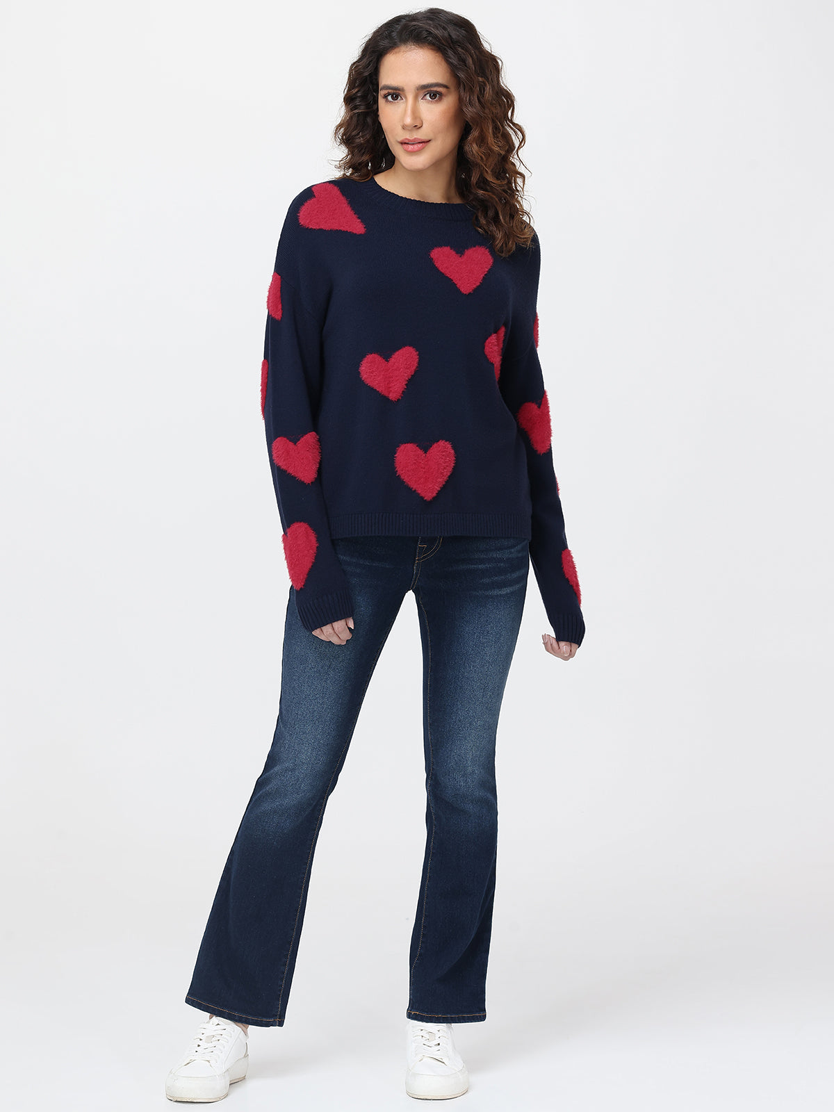Woman wearing a navy sweater with red heart patterns and blue jeans on a white background