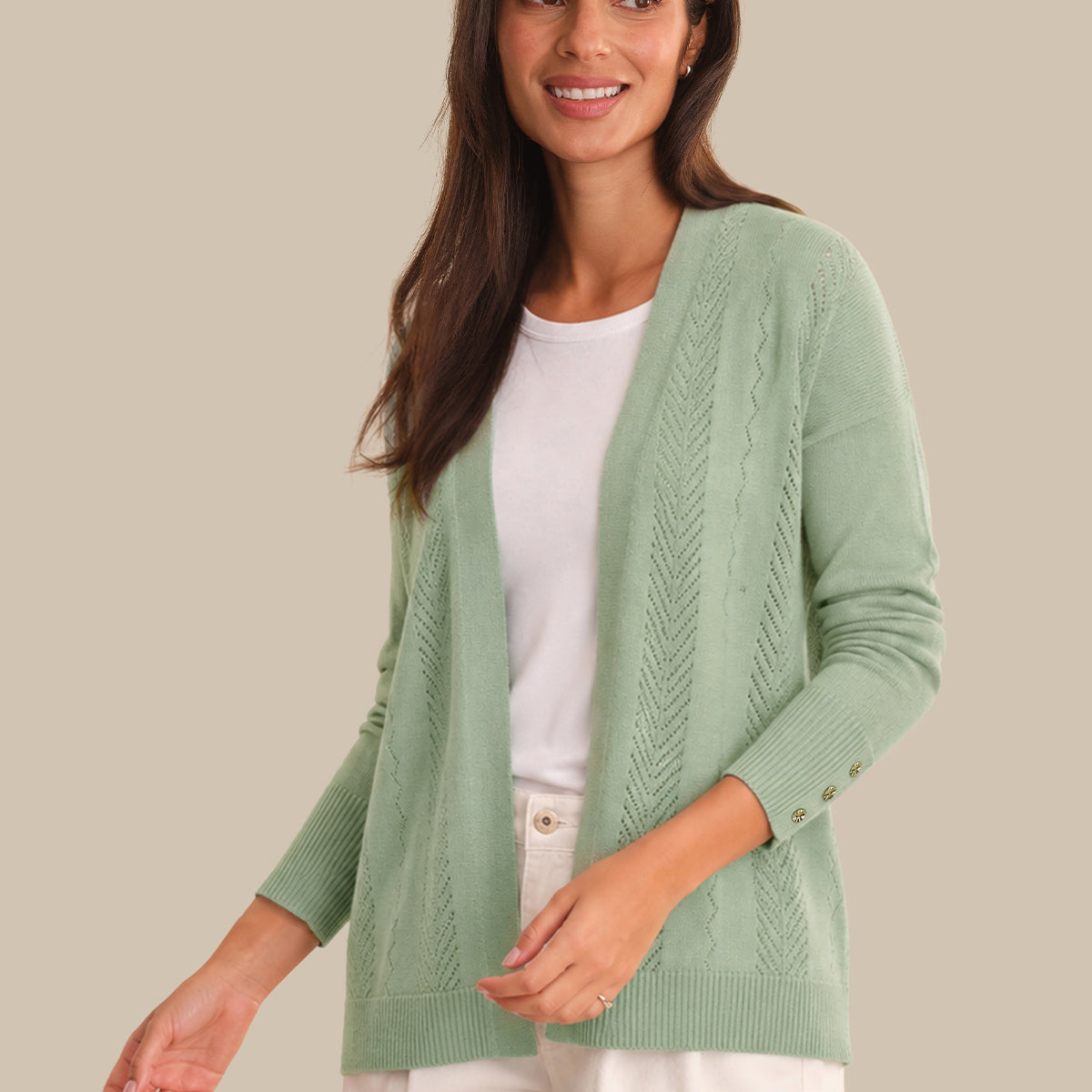 A woman wearing a sage green cable-knit open-front cardigan over a white crewneck top and white pants, standing against a solid beige background.