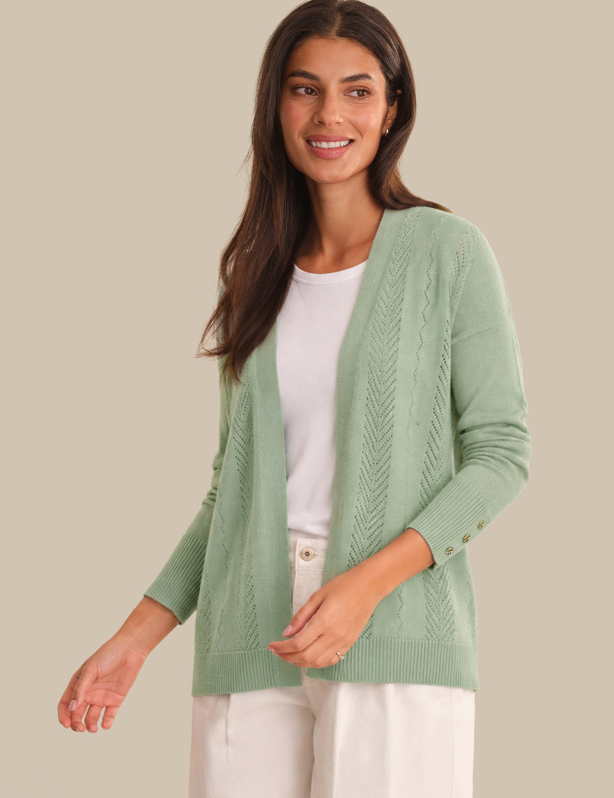 A woman wearing a sage green cable-knit open-front cardigan over a white crewneck top and white pants, standing against a solid beige background.