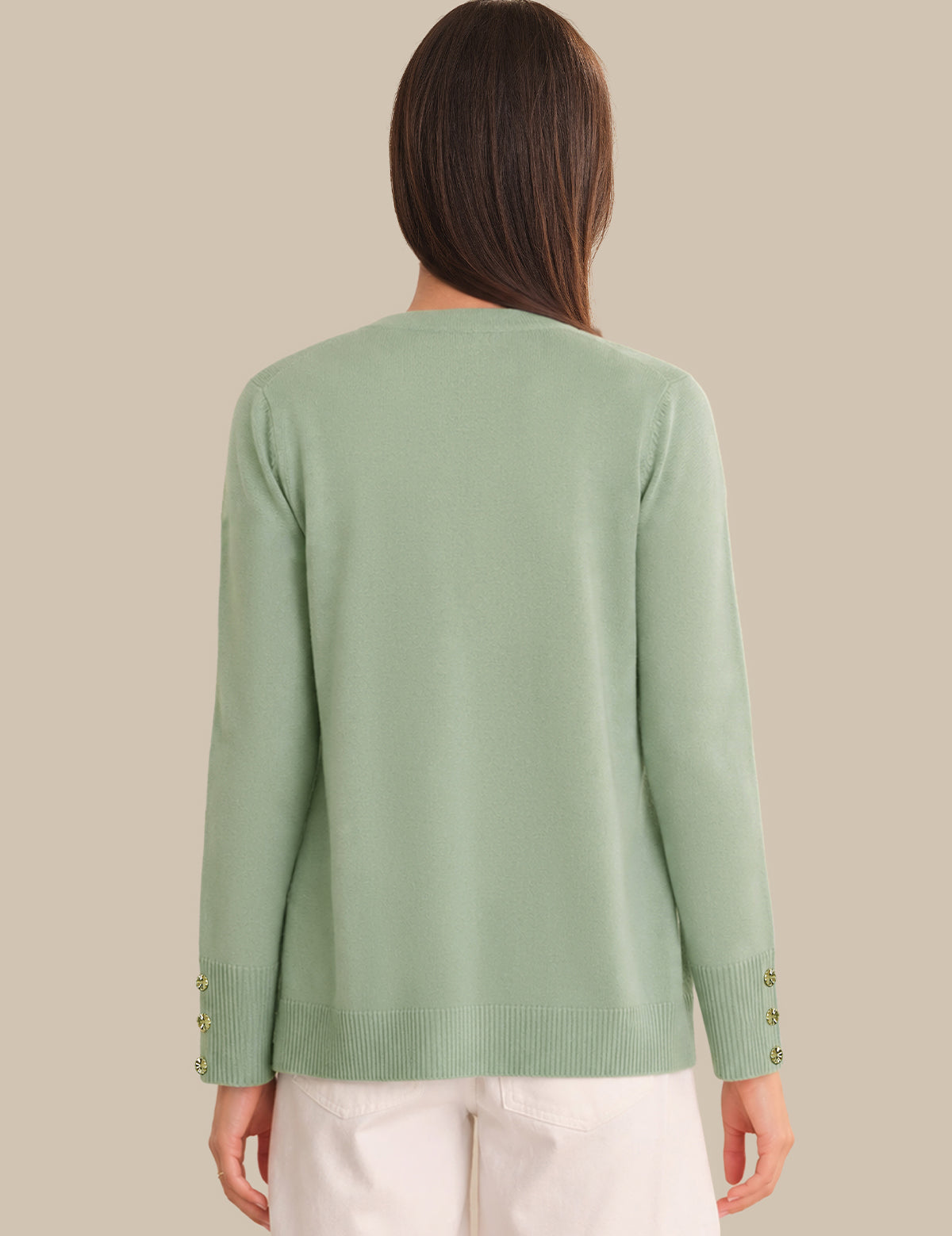 A woman viewed from the back wearing a light sage green knit sweater with decorative gold buttons on the cuffs, paired with white pants, against a solid beige background.