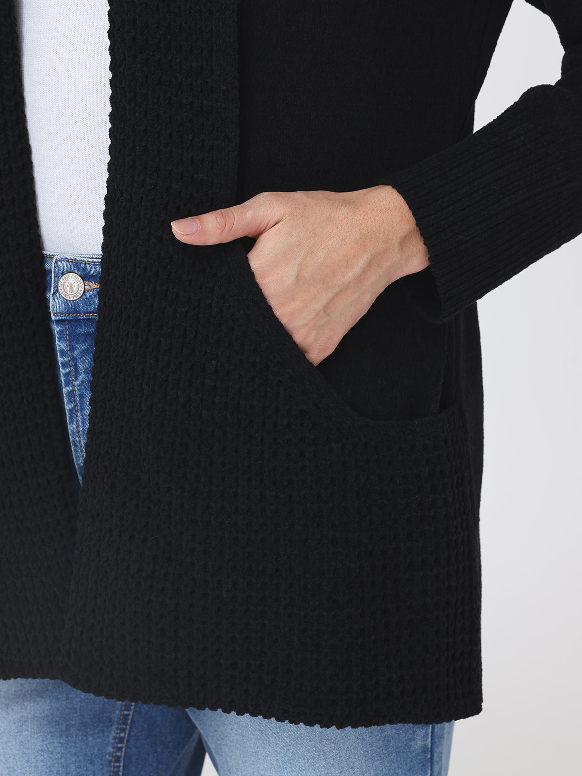 Person wearing a black knitted cardigan with a pocket, white shirt, and blue jeans on a light gray background.