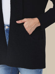 Person wearing a black cardigan with a pocket, white shirt, and blue jeans on a beige background