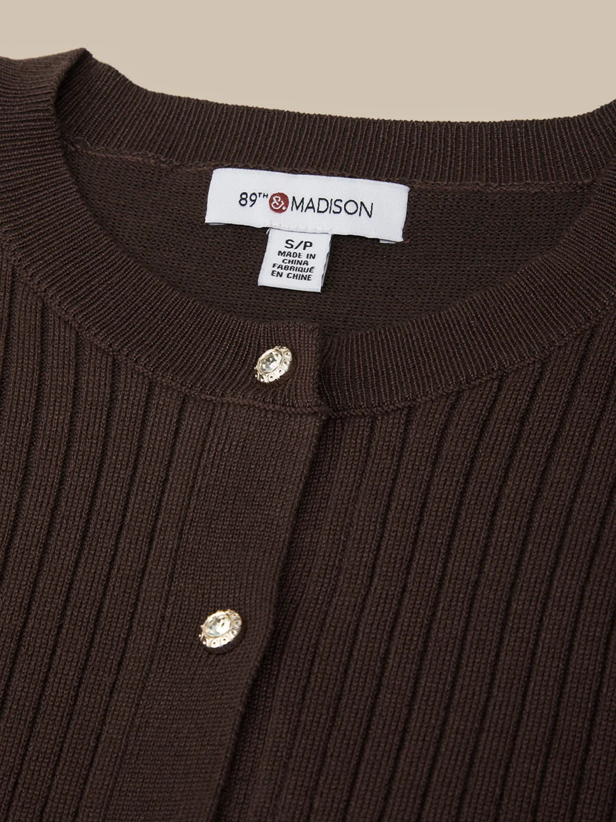 Close-up of a brown ribbed sweater with buttons and a visible label on a beige background
