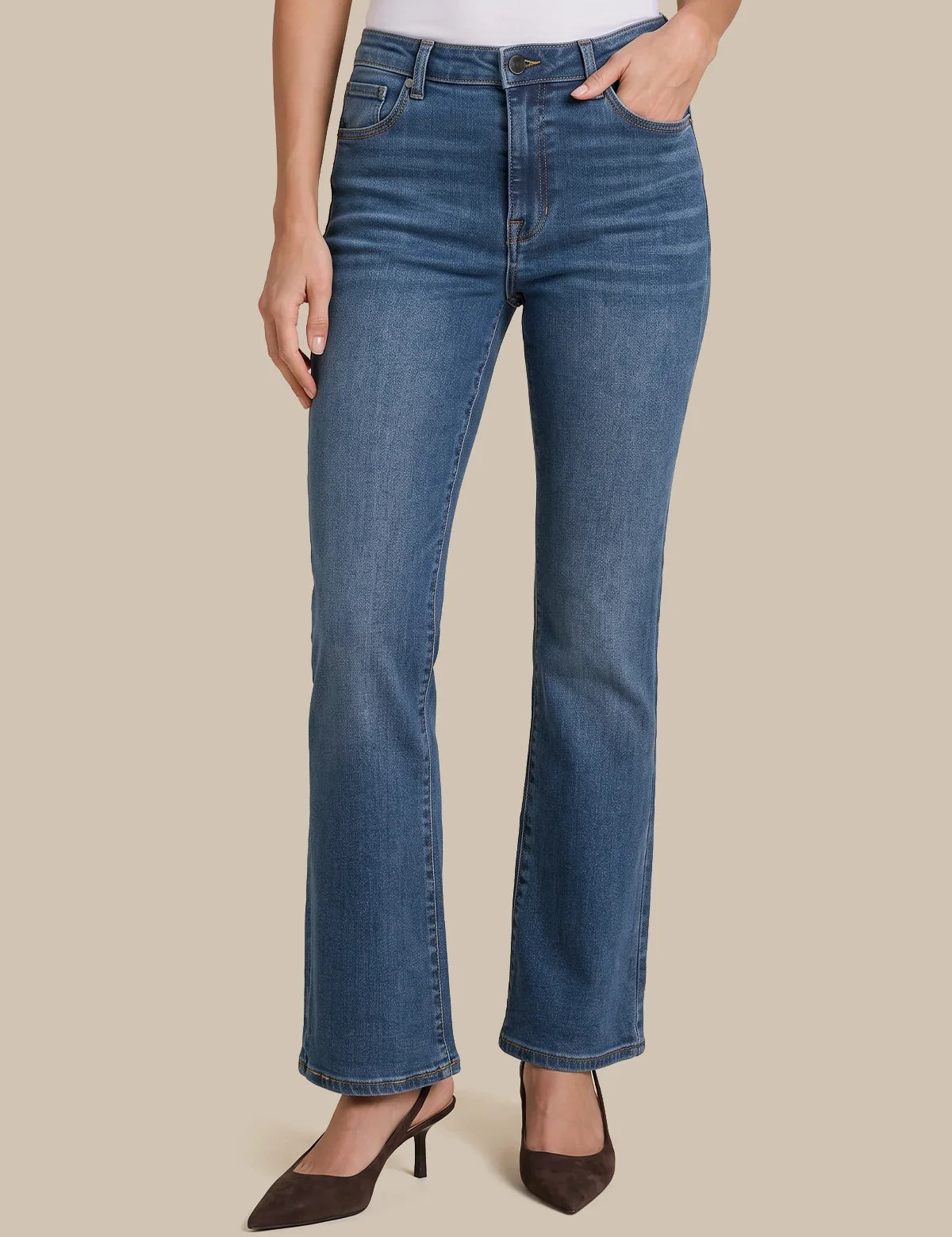 A model wearing a pair of medium-wash blue denim jeans with a straight-to-flare leg, paired with dark brown pointed-toe heels against a solid beige background.