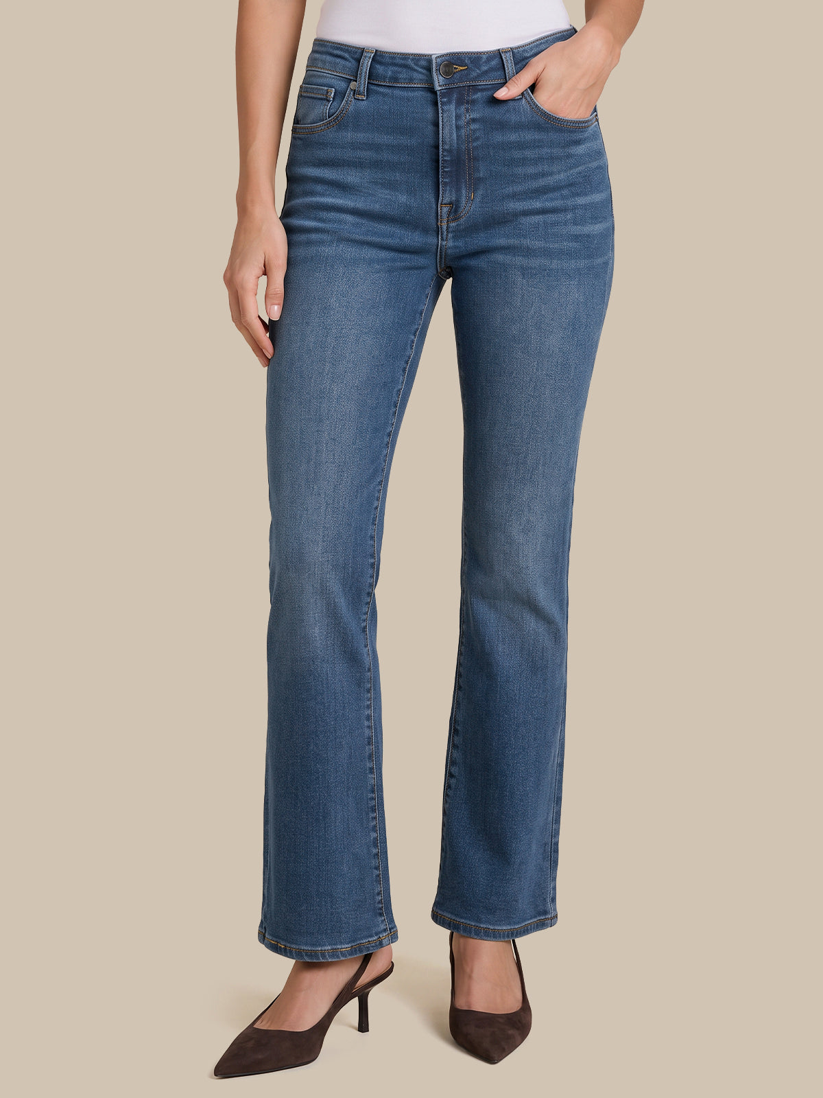 A model wearing a pair of medium-wash blue denim jeans with a straight-to-flare leg, paired with dark brown pointed-toe heels against a solid beige background.