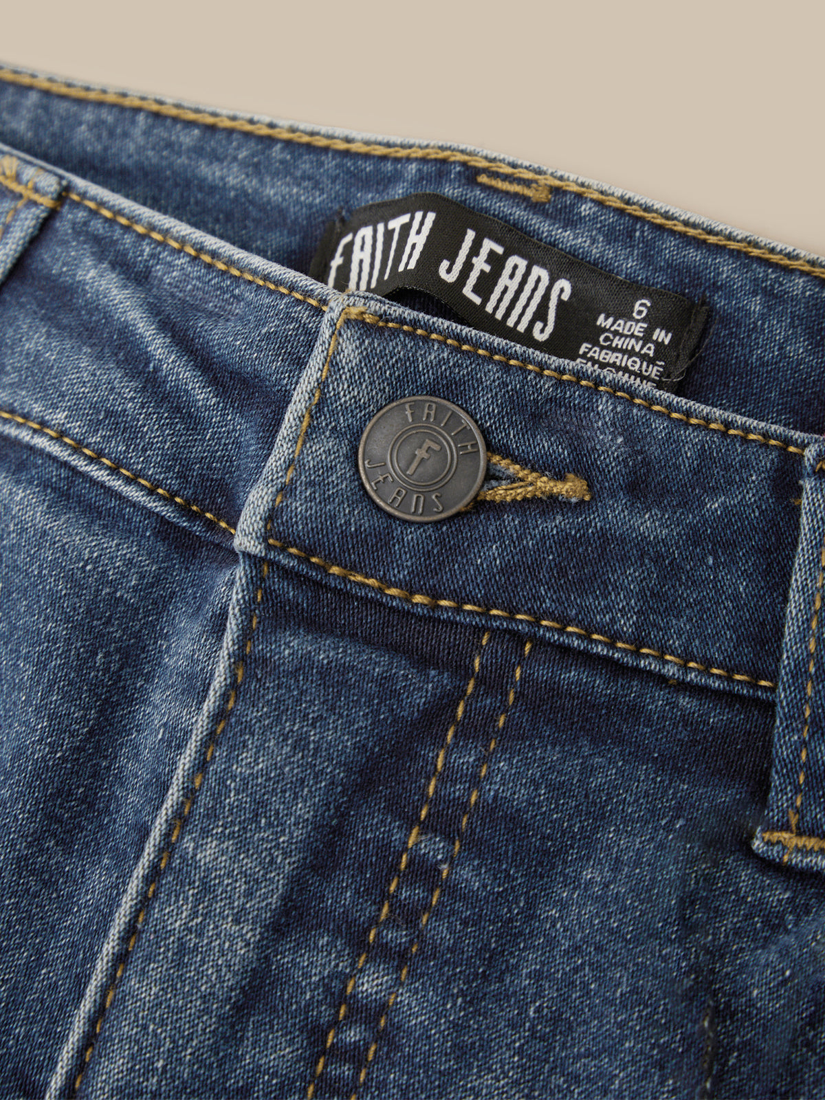 Close-up of blue denim jeans showing the waistband, black Faith Jeans brand label, metal button, and contrast stitching on a light beige background.