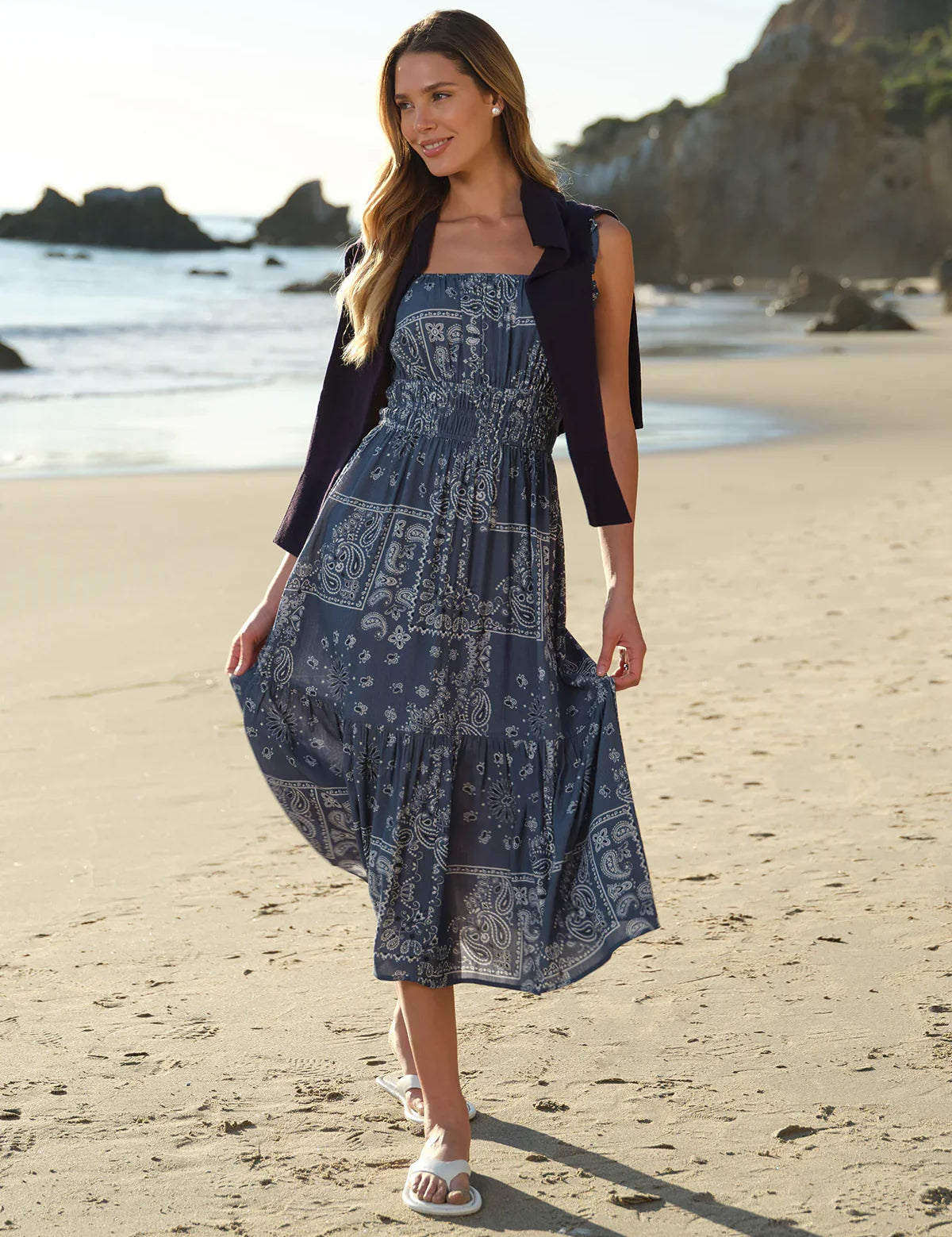 Woman in a blue patterned dress standing on a beach with ocean and rocks in the background