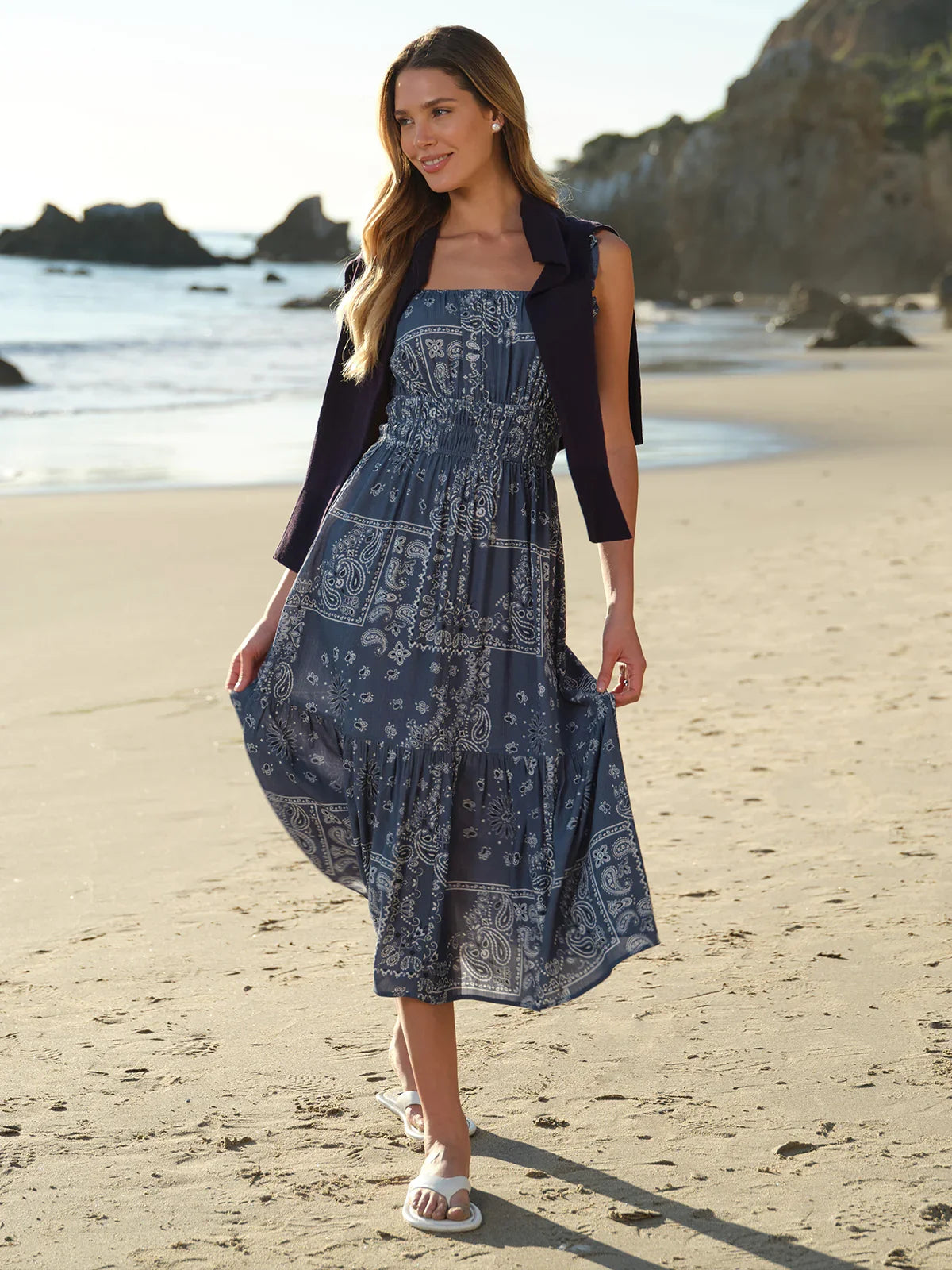Woman in a blue patterned dress standing on a beach with ocean and rocks in the background