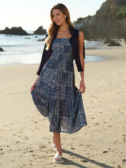 Woman in a blue patterned dress standing on a beach with ocean and rocks in the background