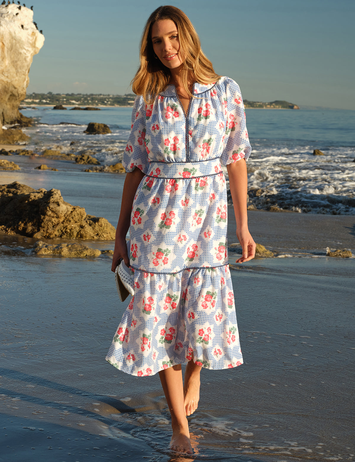 Woman in a floral dress walking on a beach with ocean and rocks in the background