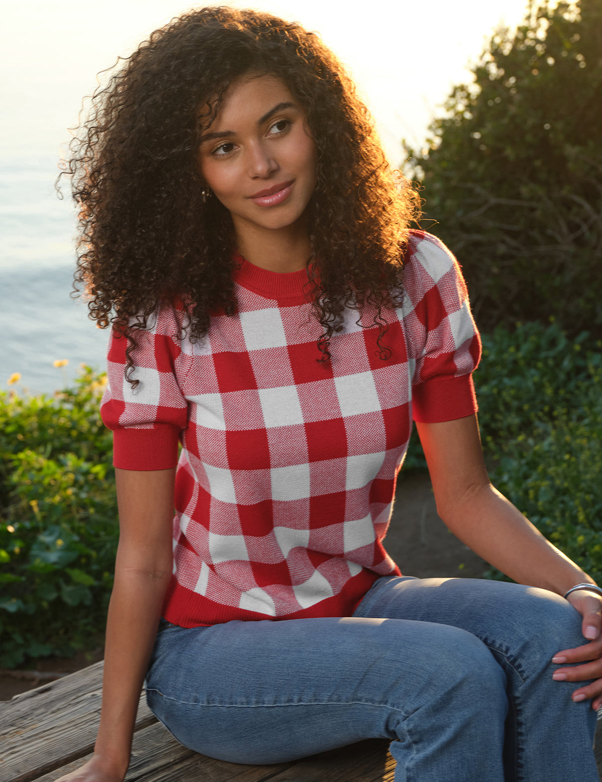 Person wearing a red and white checkered shirt sitting outdoors with greenery and water in the background