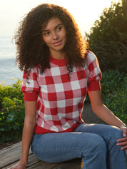 Person wearing a red and white checkered shirt sitting outdoors with greenery and water in the background