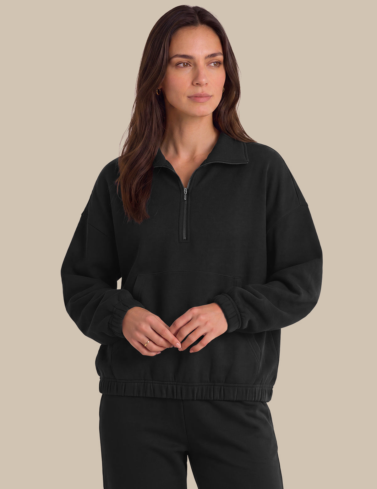 A woman modeling a black half-zip sweatshirt with a mock neck collar and matching black sweatpants, set against a solid beige background.