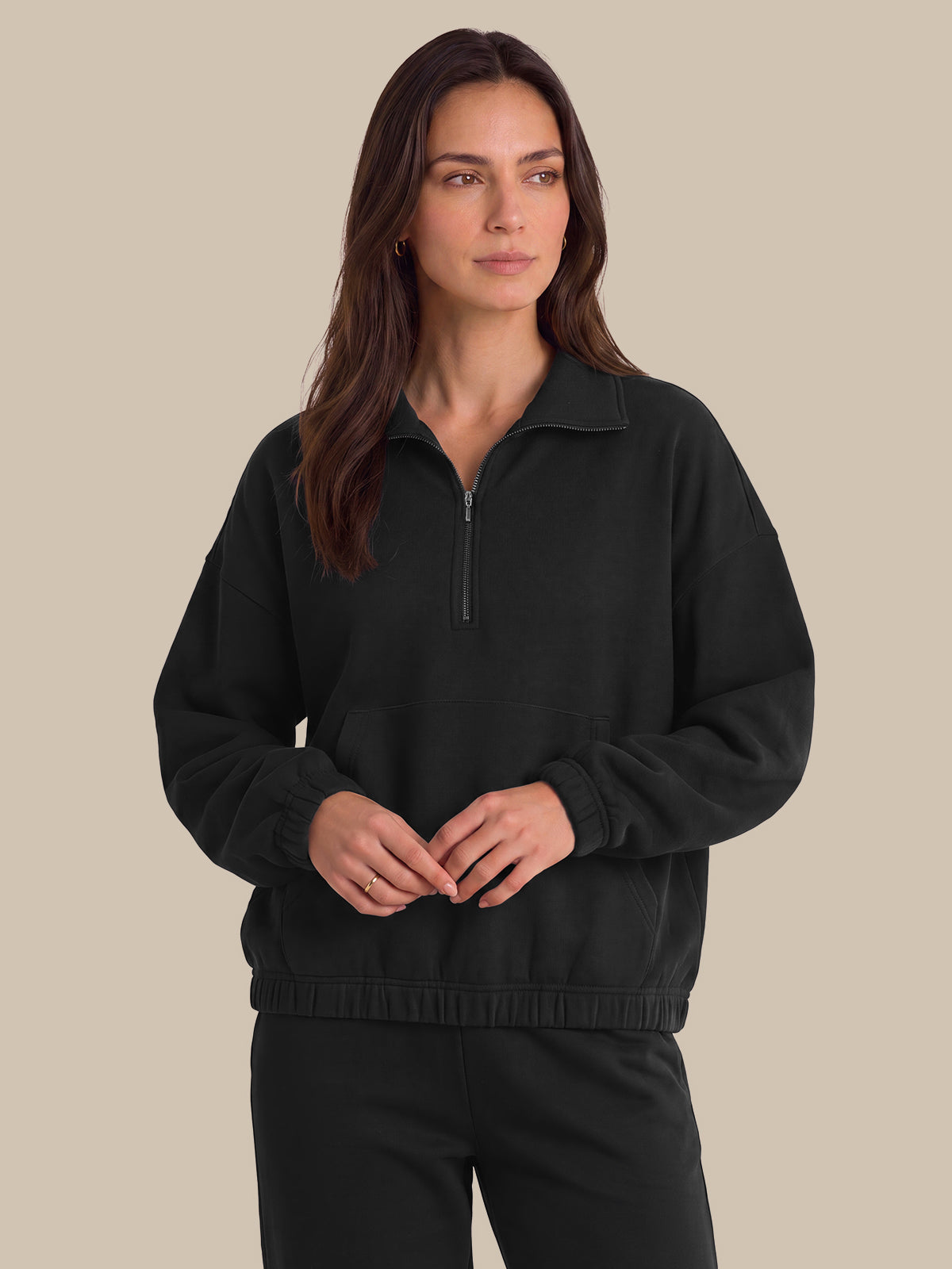 A woman modeling a black half-zip sweatshirt with a mock neck collar and matching black sweatpants, set against a solid beige background.