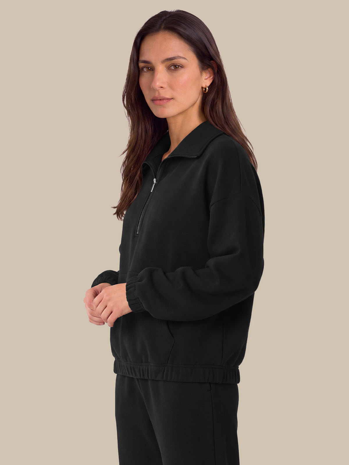 A woman modeling a black zip-up fleece jacket with a half-placket and ribbed collar, cuffs, and hem, paired with matching black pants, set against a solid beige background.
