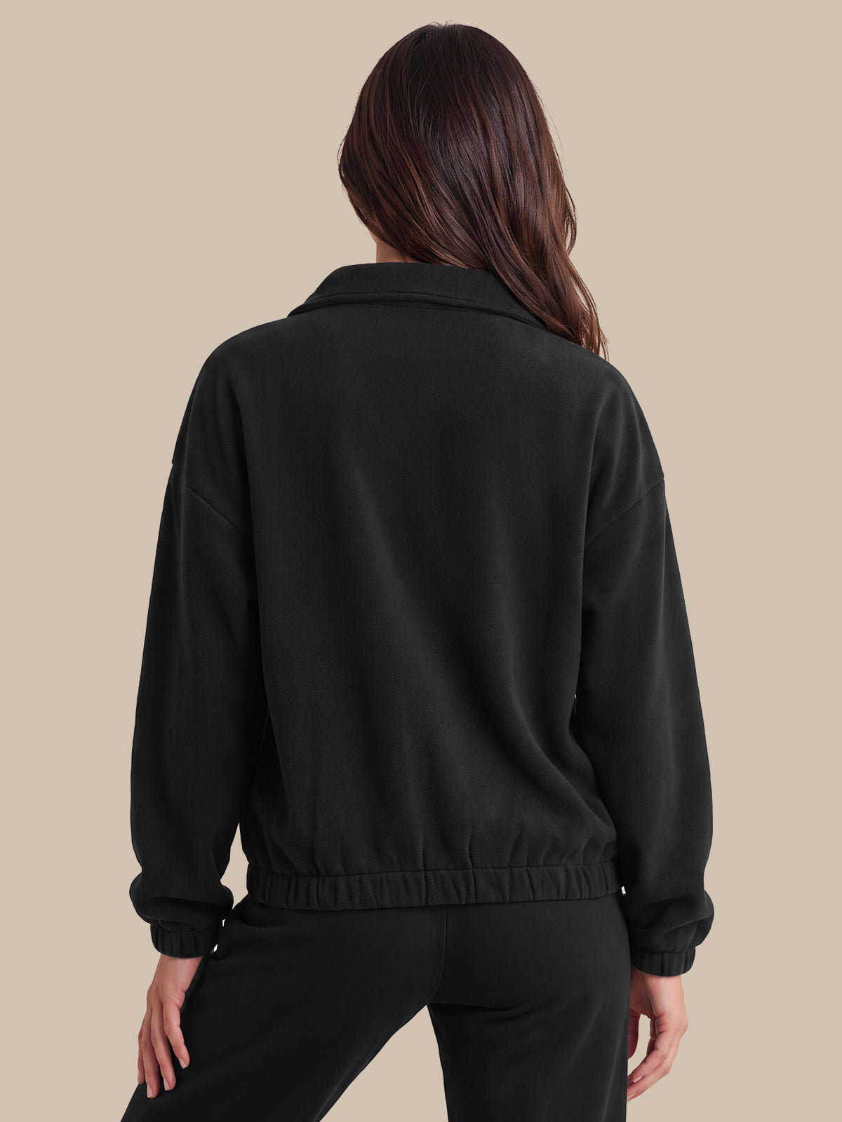 A woman shown from the back wearing a black cropped jacket with a wide collar and elasticated hem, paired with black pants, against a solid beige background.