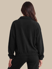 A woman shown from the back wearing a black cropped jacket with a wide collar and elasticated hem, paired with black pants, against a solid beige background.